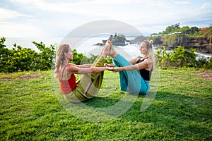 Two young women practice Ubhaya Padangusthasana, Wide-Angle Seated Forward Bend. Strengthen legs and core. Tanah Lot temple, Bali