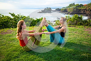 Two young women practice Ubhaya Padangusthasana, Wide-Angle Seated Forward Bend. Strengthen legs and core. Tanah Lot temple, Bali