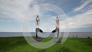 Two young women practice yoga on beach by the sea or ocea. Training tree pose.