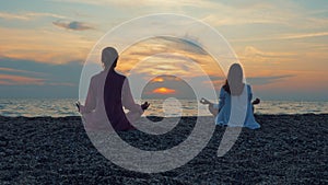 Two young women practice yoga by the sea or ocean at sunset.