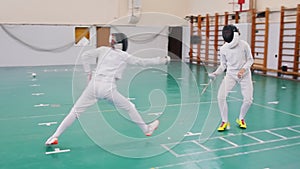 Two young women in protective helmets having an active fencing training in the school gym - making lunges on each other