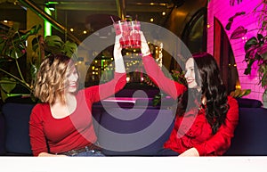 Two young women in red clothes drink cocktails and celebrate in a nightclub or bar. Women clink glasses and smyle