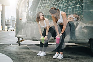 Two young women runners stand leaning against trailer, rest after training, drink water, communicate. Girls have break.