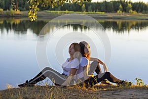Two young women sit back to back on the river bank. Friendship concept, soft focus, blur