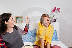 Two young women sit on the bed and cheerfully throw popcorn at each other. The concept of quarantine, work from home, security and