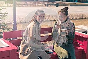 Two young women sit on a bright pink bench and drink coffee in cups, enjoying the Sunny weather and foam from coffee