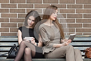 Two young women sitting on bench using their own devices