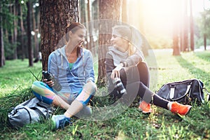 Two young women in sportswear sitting under trees in the forest drinking water, talking and resting after training