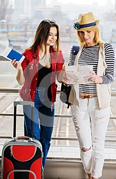 Two young women stand with a suitcase at the railway station or the airport. Look at the card and passport.