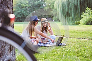 Two young women students in park sitting on grass talking, using laptop