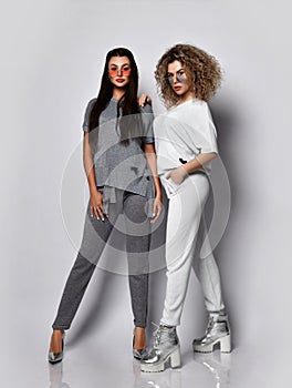 Two young women in stylish glasses, white and gray suits and silver footwear pose together standing walking