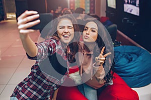 Two young women taking selfie on phone camera and posing in playing room. They shink and show fingers. Cheerful models