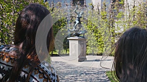 Two young women walking in the park among decorative plants and taking pictures of statues