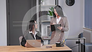 Two young women are working in the office. A woman is talking on the phone, and her colleague is making notes in a notebook