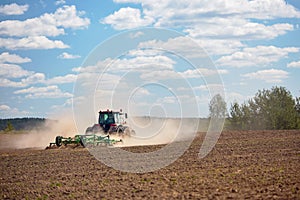 Typical agricultural scene tractor cultivation in field in clouds of dust drives off into the distance with birches