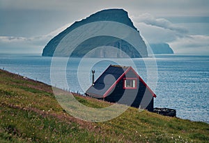 Typical Faroese black walls house and Koltur island on background. Gloomy summer scene of outskirts of Sandavagur village,  Vagar