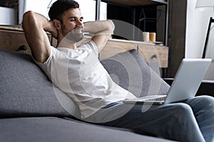 Typing new blog post. Side view of handsome young man using his laptop with smile while sitting on the couch at home.