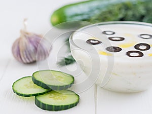 Tzatziki in a glass bowl with cucumber slices and dill, and garlic on a rustic white wooden table.