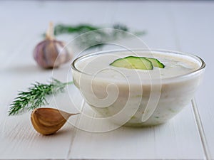 Tzatziki in a glass bowl with cucumber slices and dill, and garlic on a white wooden table.