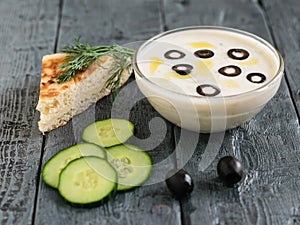 Tzatziki sauce in a glass bowl and olives on a dark wooden table with a piece of bread.