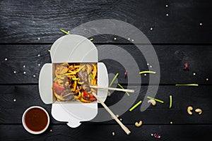 Udon stir fry noodles with meat and vegetables in a box on black background. With chopsticks and sauce.