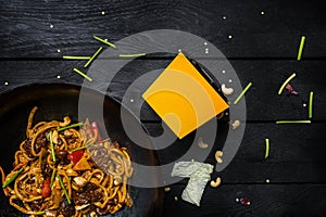 Udon stir fry noodles with meat and vegetables in wok pan on black wooden background. With a box for noodles