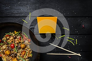 Udon stir fry noodles with seafood and vegetables in wok pan on black wooden background. With a box for noodles