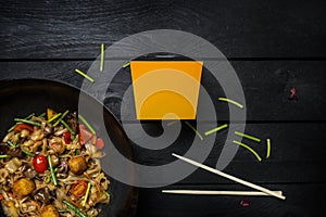 Udon stir fry noodles with seafood and vegetables in wok pan on black wooden background. With a box for noodles