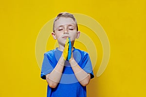 Ukrainian boy prays for Ukraine. Children against war. A boy in a blue T-shirt on a yellow background folded his hands painted in