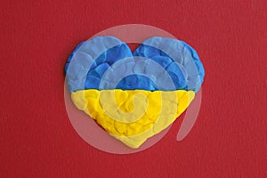 Ukrainian flag. Heart shape of blue and yellow plasticine modeling clay on red maroon texture background. Plasticine finger