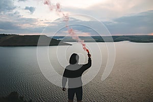 Ultras hooligan holding red smoke bomb in hand, standing on top of rock mountain with amazing sunset view on river. Atmospheric