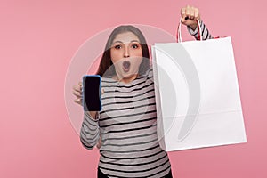 Unbelievable online store app! Portrait of shocked woman in sweatshirt holding shopping bag and cell phone