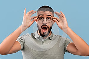 Unbelievable. Shocked middle eastern man touching his glasses and opening mouth, looking at camera on blue background