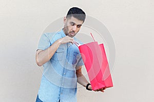 Unboxing and looking inside of bag. Portrait of handsome young bearded man in blue shirt standing, holding red shopping bag,