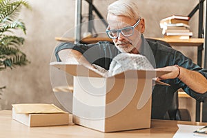 Unboxing parcel. Senior man looking into a cardboard box at home, delivery order from an online store