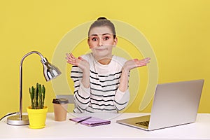 Uncertain woman office worker in striped shirt shrugging shoulders sitting on laptop at workplace.