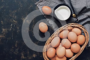 Uncooked brown eggs in a basket and milk in a jug on a dark rustic background. Top view, copy space.