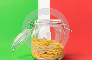 Uncooked penne pasta in glass crystal storage jar with open lid on white green red background flag colors. Italian cuisine