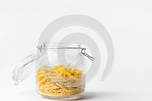 Uncooked penne pasta in glass crystal storage jar with open lid on white table wall background. Italian cuisine specialty. Kitchen