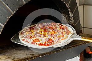 Uncooked salami pizza on metal paddle in front of wood oven.
