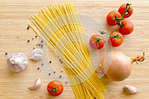 Uncooked spaghetti with garlic cloves, cherry tomatoes, onion and peppercorns on a cutting board.