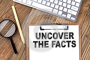 UNCOVER THE FACTS text on paper clipboard with magnifier and keyboard on wooden background