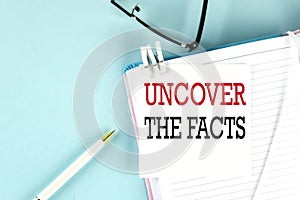 UNCOVER THE FACTS text on a sticky on notebook with pen and glasses , blue background