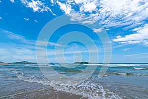 Under the blue sky and white clouds, the waves wash on the beach, and the small island in the distance