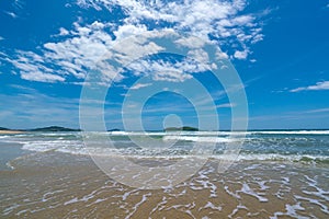 Under the blue sky and white clouds, the waves wash on the beach, and the small island in the distance