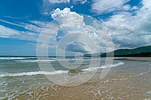 Under the blue sky and white clouds, the waves wash on the beach, and the small island in the distance