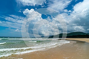 Under the blue sky and white clouds, the waves wash on the beach, and the small island in the distance