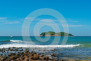 Under the blue sky and white clouds, the waves wash on the beach, and the small island in the distance