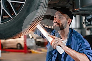 Under car, using lighting device. Auto mechanic working in garage. Repair service