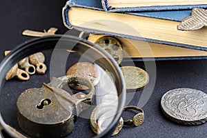 Under a magnifying glass is considered an old copper padlock around an old coin and a book, a dark cloth background